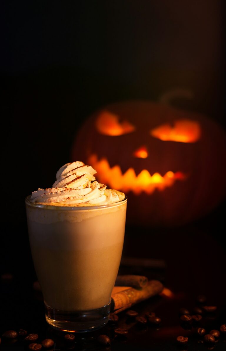 A tall glass of pumpkin spice latte under whipped cream dusted with spice, cinnamon sticks and coffee beans beside it, a lit jack-o'-lantern behind