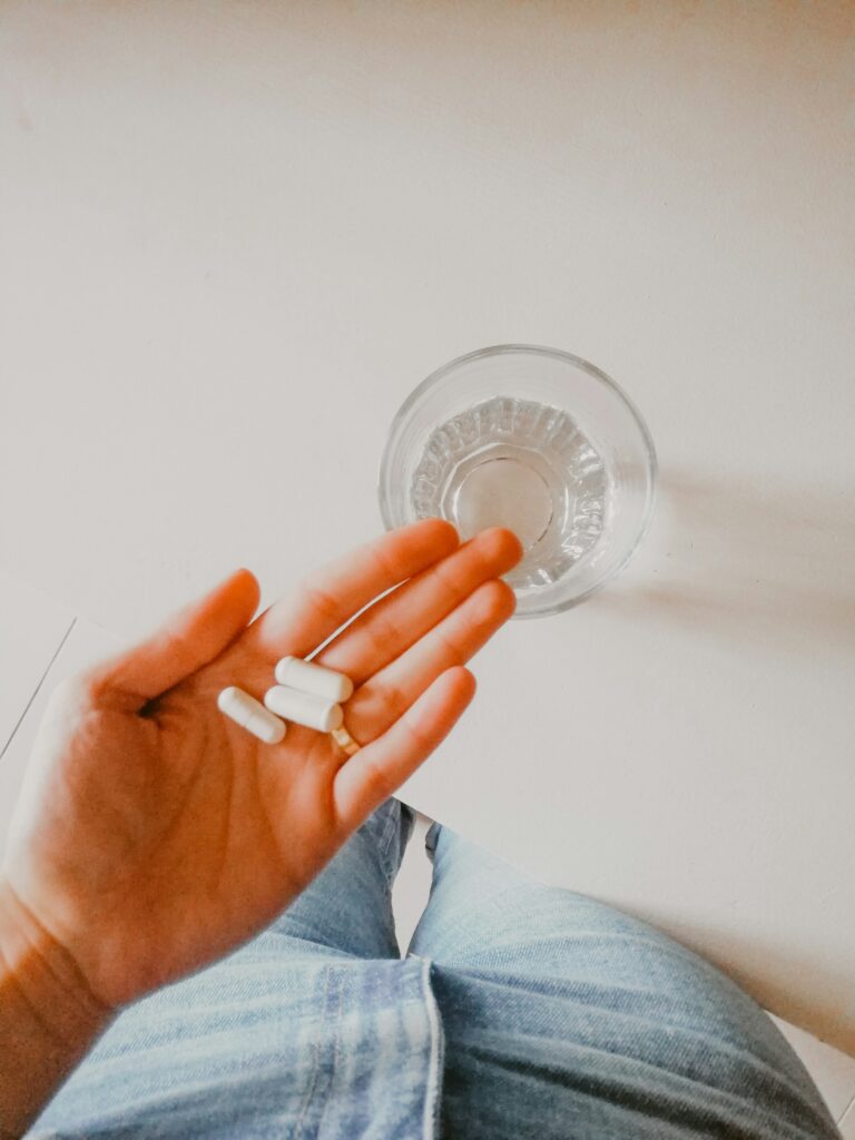 A hand holding white capsules above a glass of water