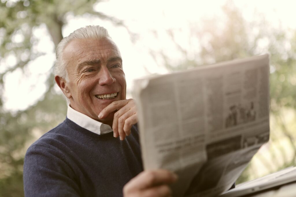 A gray-haired older man smiling as he reads a newspaper outdoors